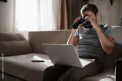 Funny man looking searching binoculars a laptop on the table working at home office.