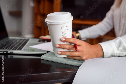 Young business woman sitting in cafe and taking notes in notebook. On table is laptop and a paper cup of coffee. Student learning online. Blogger.