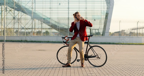 Male casual handsome bike rider in red jacket sitting on bicycle, sipping hot drink and talking on cellphone. At big glass modern building. Caucasian man drinking coffee. and speaking on mobile phone.