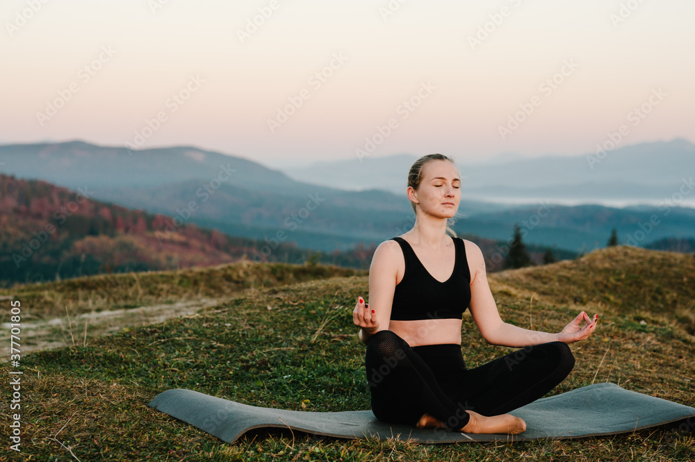 A young woman is practicing yoga at mountains. Relaxing in nature. Young girl doing yoga fitness exercise outdoor in the beautiful landscape. Morning sunrise, Namaste Lotus pose. Meditation.