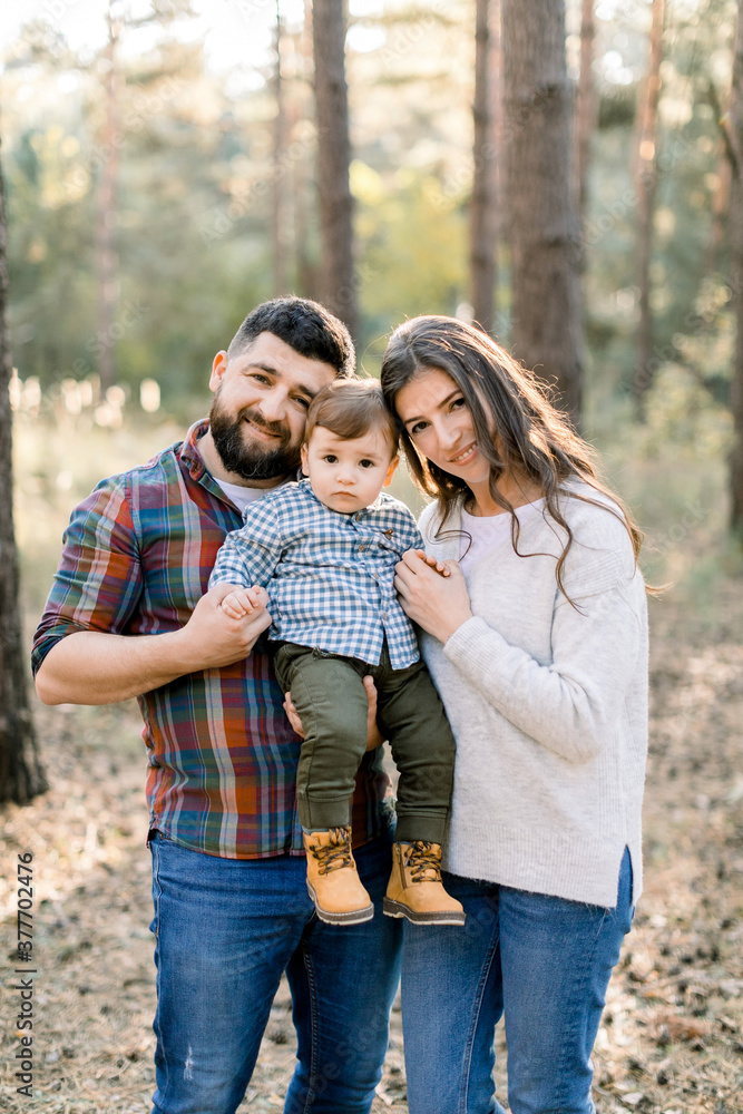 Happy family, dad, mom and little son, wearing stylish casual clothes, walking in the autumn pine forest at beautiful sunny day. Joyful parents holding little baby son and smiling to camera Happy family, dad, mom and little son, wearing stylish casual clothes, walking in the autumn pine forest at beautiful sunny day. Joyful parents holding little baby son and smiling to camera
