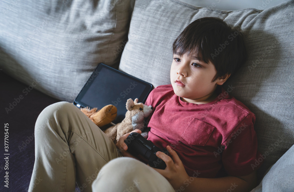 Portrait of kid holding video game or game console. Child playing game online at home, Young boy siting alone on sofa relaxing on his own on weekend, New normal lifestlye concept Portrait of kid holding video game or game console. Child playing game online at home, Young boy siting alone on sofa relaxing on his own on weekend, New normal lifestlye concept