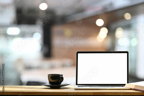 White blank screen laptop, black coffee cup on the wooden working desk over blurred modern office background.