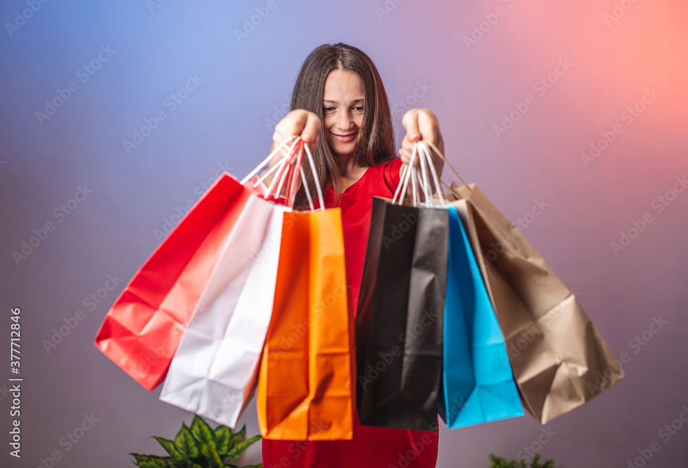 Young woman in a red dress is holding a lot of multicolored paper bags with purchases. Concept of shopping and sales. Blue and pink background