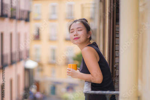 young beautiful and happy Asian Korean woman enjoying city view from hotel room balcony in Spain during holidays trip in Europe drinking coffee relaxed in urban background