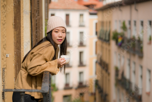 young beautiful and happy Asian Korean woman in winter hat enjoying city view from hotel room balcony in Spain during holidays trip in Europe drinking coffee relaxed