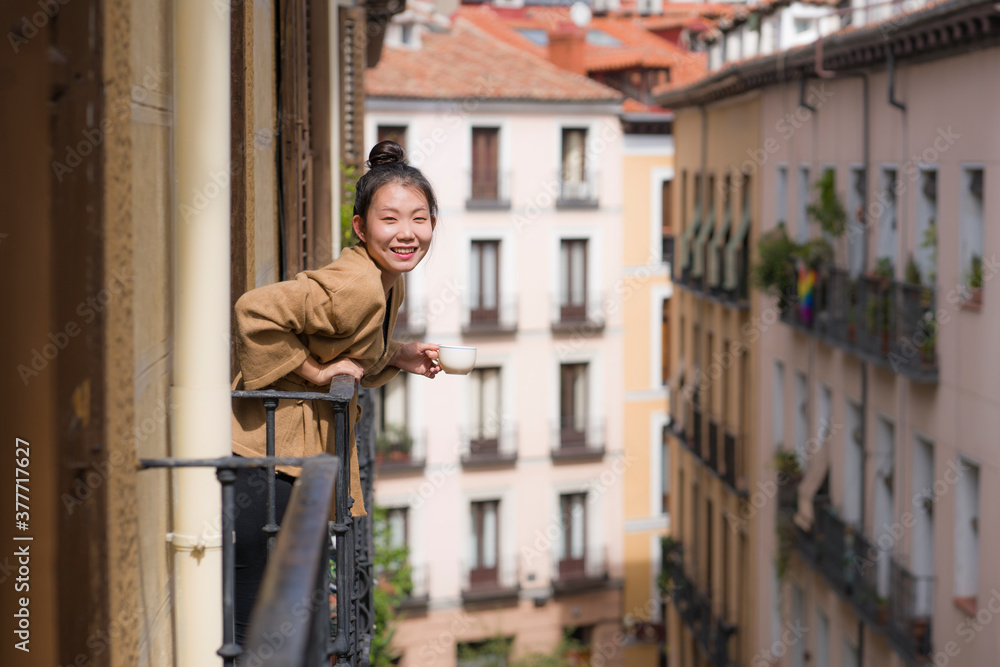 young happy and beautiful Asian Chinese woman enjoying city view from hotel room balcony in Spain during holidays trip in Europe drinking coffee smiling cheerful