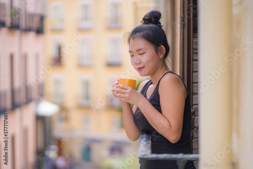 young beautiful and happy Asian Chinese woman enjoying city view from hotel room balcony in Spain during holidays trip in Europe drinking coffee relaxed in urban background