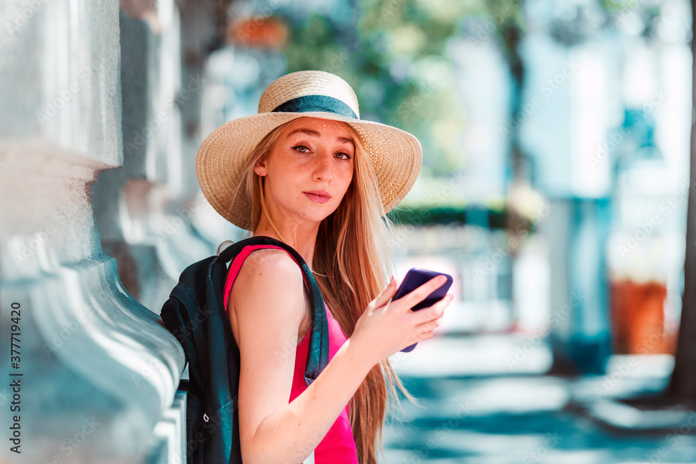Happy young woman wearing straw hat while standing on the street and text messaging