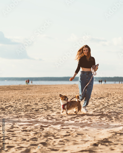 Beautiful young woman and Corgi puppy running along beach by water. Female walking with dog on Sunny day on ocean coastline. Active lifestyle, pet care