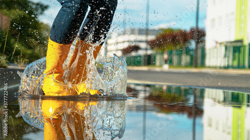 LOW ANGLE: Girl in bright yellow rubber boots jumps into the glassy puddle. photo