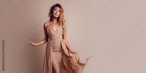 Gorgeous  seductive woman with blonde wavy hairs posing in studio. wearing elegant beige sequins  dress. Space for text. Beige background.
