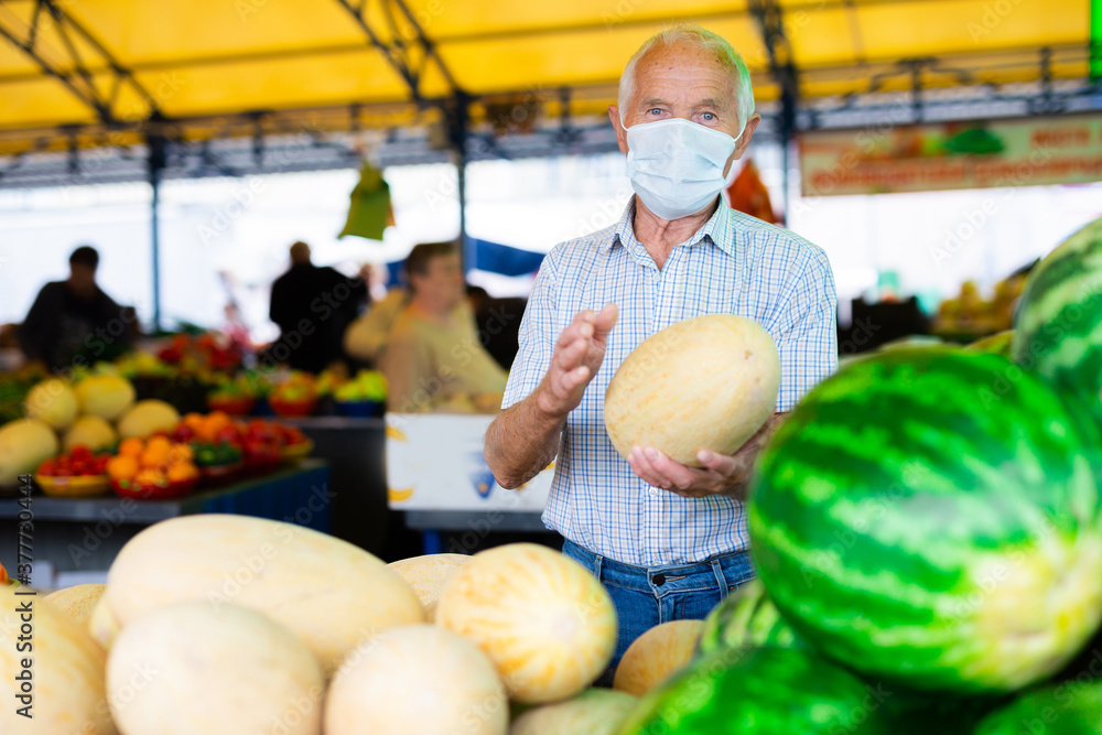 Fototapeta premium retired european man wearing medical mask protecting against the virus selling melons and watermelons in market