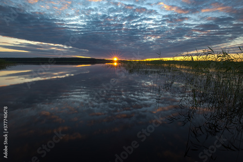 The sun with bright beams rises over the lake. The sky with lightened clouds is reflected in the mirror-like water
