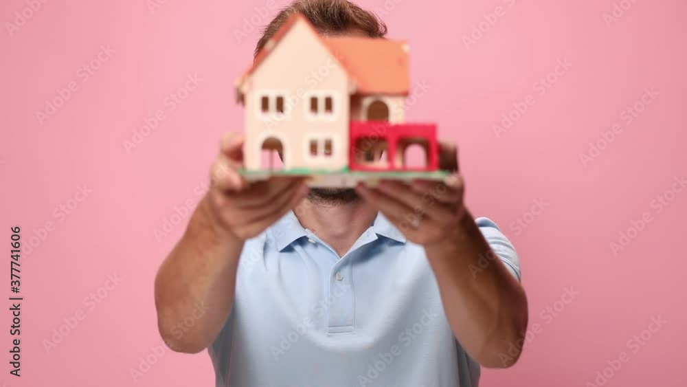 smiling young casual in blue polo shirt holding house model in front of face and presenting on pink background smiling young casual in blue polo shirt holding house model in front of face and presenting on pink background