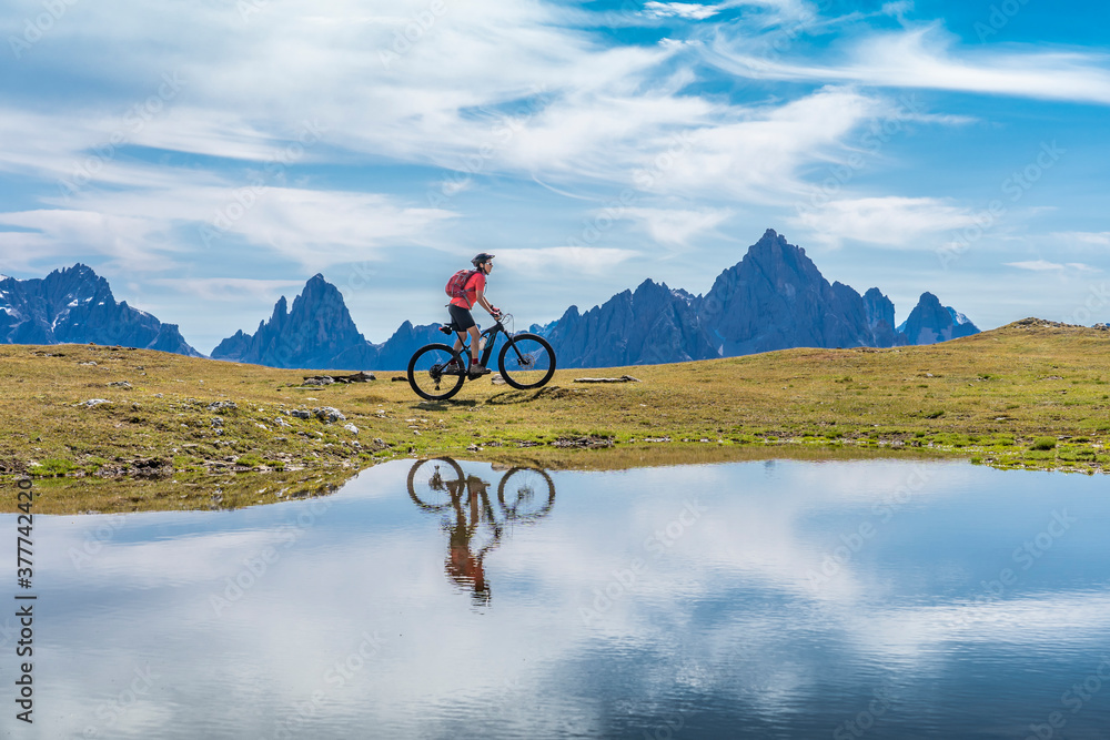 nice woman riding her electric mountain bike the Three Peaks Dolomites, reflecting herself in the blue water of a cold mountain lake