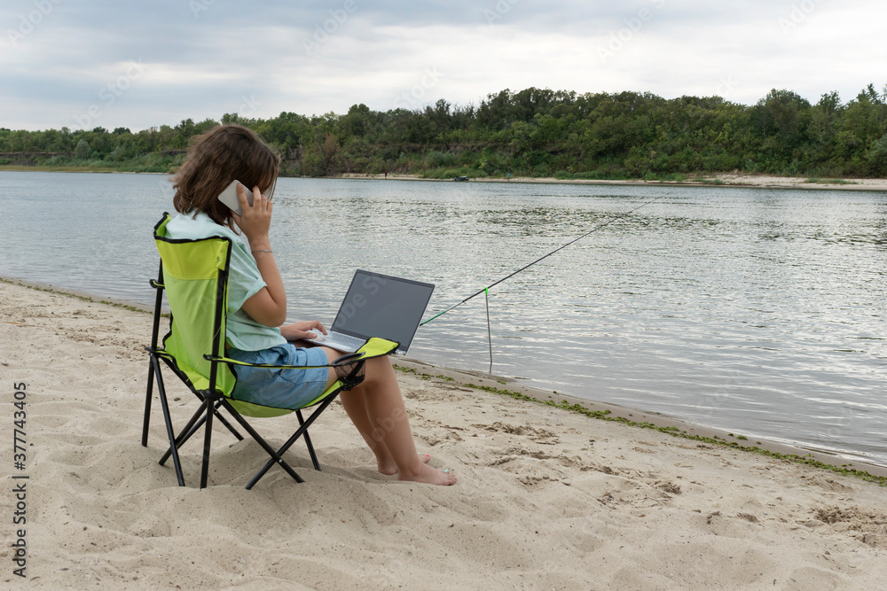 young female sit on bank of summer river with laptop on her knees. Remote work concept. Opportunity to be in touch on distance.