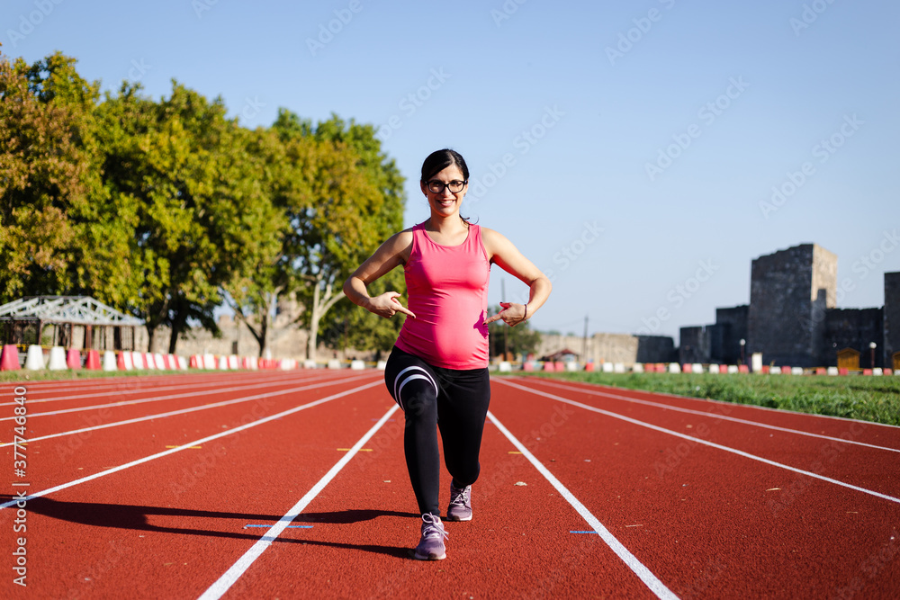 Active, 6 months pregnant woman workingout on track field on sunny morning. Attractive female doing exercise outdoors and proudly pointing on her belly. Active, 6 months pregnant woman workingout on track field on sunny morning. Attractive female doing exercise outdoors and proudly pointing on her belly.