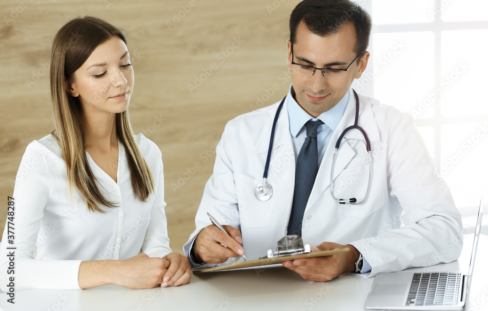 Doctor and patient discussing medical exam results while sitting at the desk in sunny clinic Doctor and patient discussing medical exam results while sitting at the desk in sunny clinic