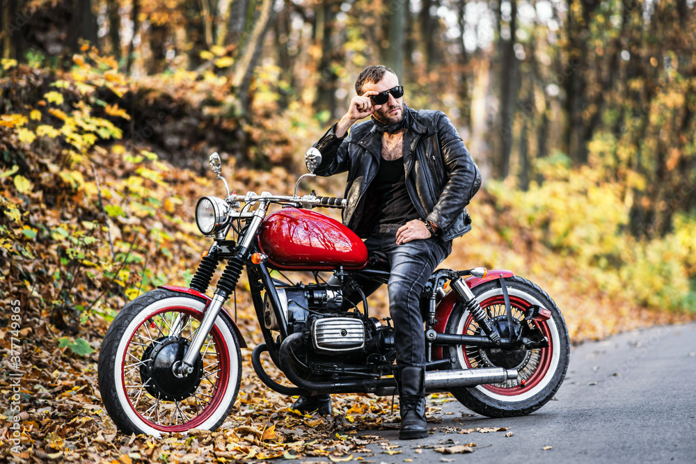 Fototapeta premium Bearded brutal man in sunglasses and leather jacket sitting on a motorcycle on the road in the forest