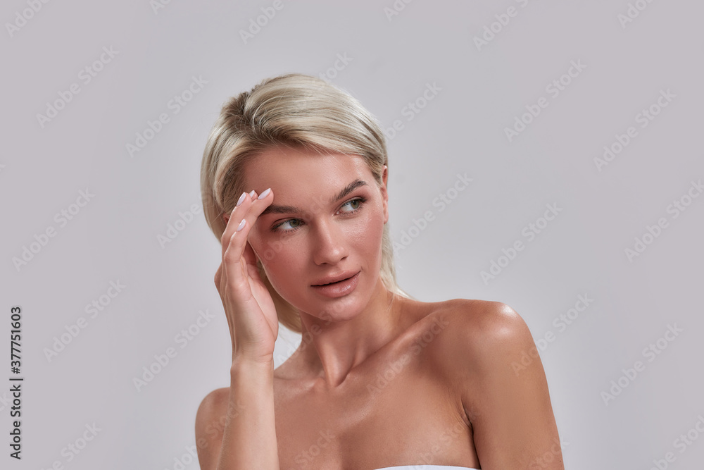 Portrait of beautiful young female blonde with perfect fresh skin looking aside, touching her face while posing isolated over grey background Portrait of beautiful young female blonde with perfect fresh skin looking aside, touching her face while posing isolated over grey background
