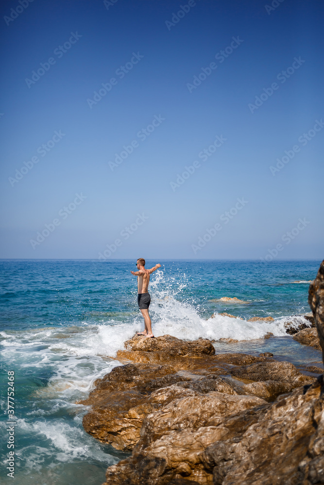 A young man stands on the rocks overlooking the open Mediterranean Sea. A guy on a warm summer sunny day looks at the sea breeze