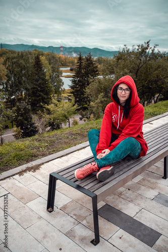 A female student in a red jacket sits on a bench in a park. Against the backdrop of a large autumn forest in cloudy weather