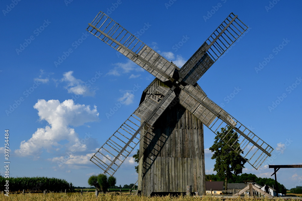 View of a massive wooden historic windmill with its body and blades made out of planks, logs, and boards standing in the middle of a field ready to be harvested seen on a cloudy summer day in Poland