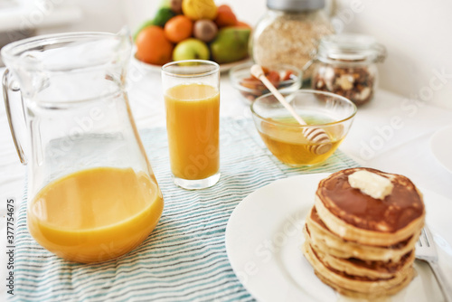 A jug and a glass of orange juice, honey in a bowl, a stack of pancakes and various fruits served for breakfast on the table