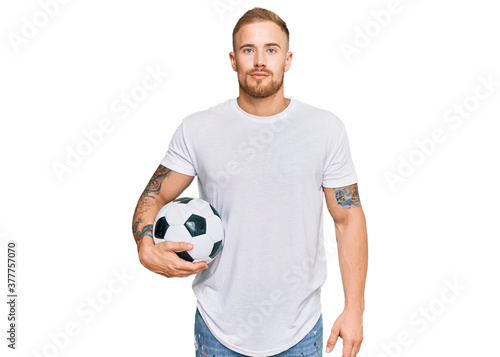 Young irish man holding soccer ball thinking attitude and sober expression looking self confident