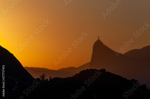 Beautiful distant view of Christ the Redeemer silhouette at sunset against orange sky; Mount Corcovado, Rio de Janeiro, Brazil. Smaller mountain in the foreground. Copy space for text.