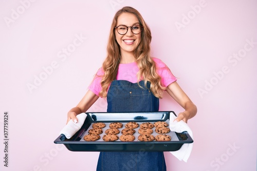 Fotografia Young caucasian woman with blond hair wearing baker uniform holding homemade cookies winking looking at the camera with sexy expression, cheerful and happy face
