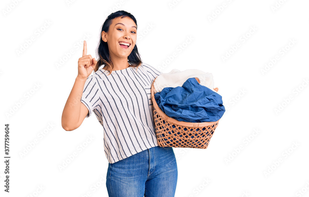Young woman holding laundry basket surprised with an idea or question pointing finger with happy face, number one Young woman holding laundry basket surprised with an idea or question pointing finger with happy face, number one
