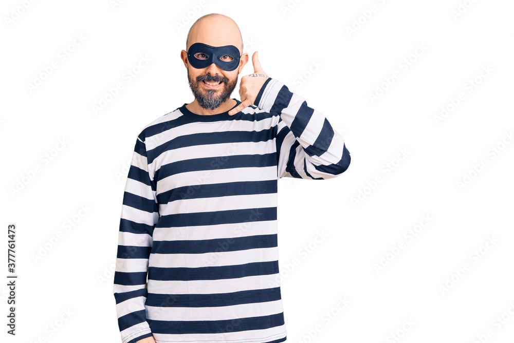 Young handsome man wearing burglar mask smiling doing phone gesture with hand and fingers like talking on the telephone. communicating concepts. Young handsome man wearing burglar mask smiling doing phone gesture with hand and fingers like talking on the telephone. communicating concepts.