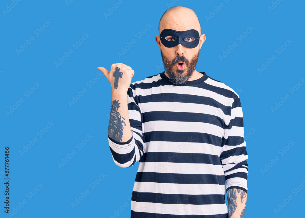 Naklejka premium Young handsome man wearing burglar mask surprised pointing with hand finger to the side, open mouth amazed expression.