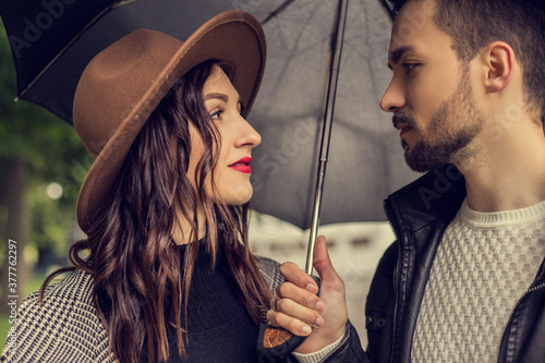 Portrait of young loving couple walking in the city park and use umbrella to shelter the rain. Heterosexual lovers have a date, spending time together in cloudy rainy day, showing emotions.