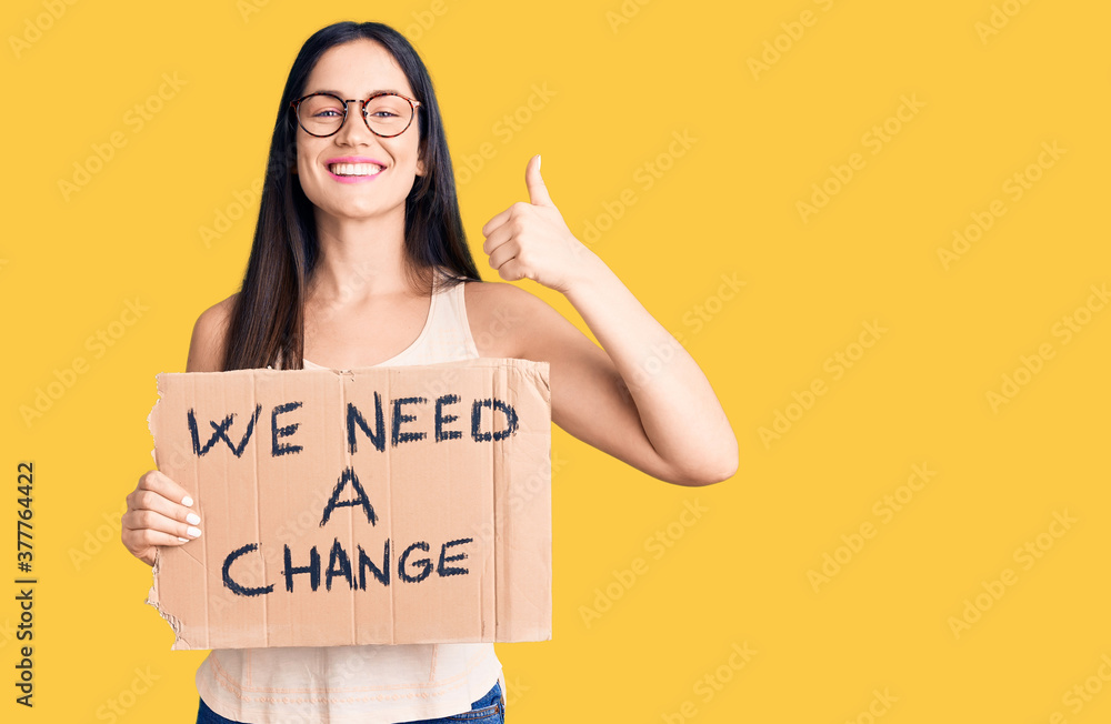 Young beautiful caucasian woman holding we need a change banner smiling happy and positive, thumb up doing excellent and approval sign Young beautiful caucasian woman holding we need a change banner smiling happy and positive, thumb up doing excellent and approval sign