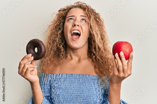 Beautiful caucasian teenager girl holding red apple and donut angry and mad screaming frustrated and furious  shouting with anger looking up.
