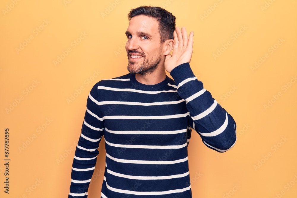 Young handsome man wearing casual striped sweater standing over isolated yellow background smiling with hand over ear listening and hearing to rumor or gossip. Deafness concept. Young handsome man wearing casual striped sweater standing over isolated yellow background smiling with hand over ear listening and hearing to rumor or gossip. Deafness concept.