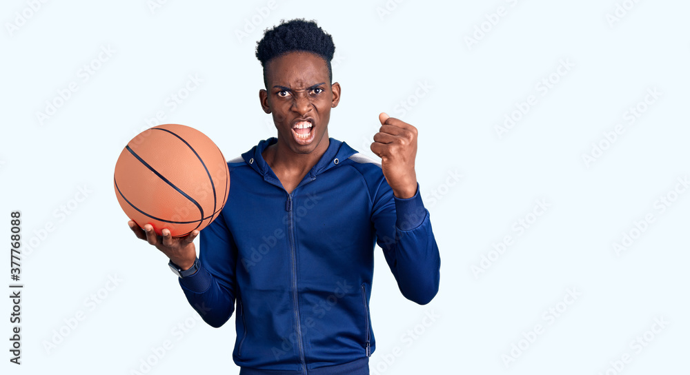 Young african american man holding basketball ball annoyed and frustrated shouting with anger, yelling crazy with anger and hand raised