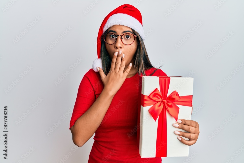 Young latin girl wearing christmas hat and holding a gift covering mouth with hand, shocked and afraid for mistake. surprised expression Young latin girl wearing christmas hat and holding a gift covering mouth with hand, shocked and afraid for mistake. surprised expression