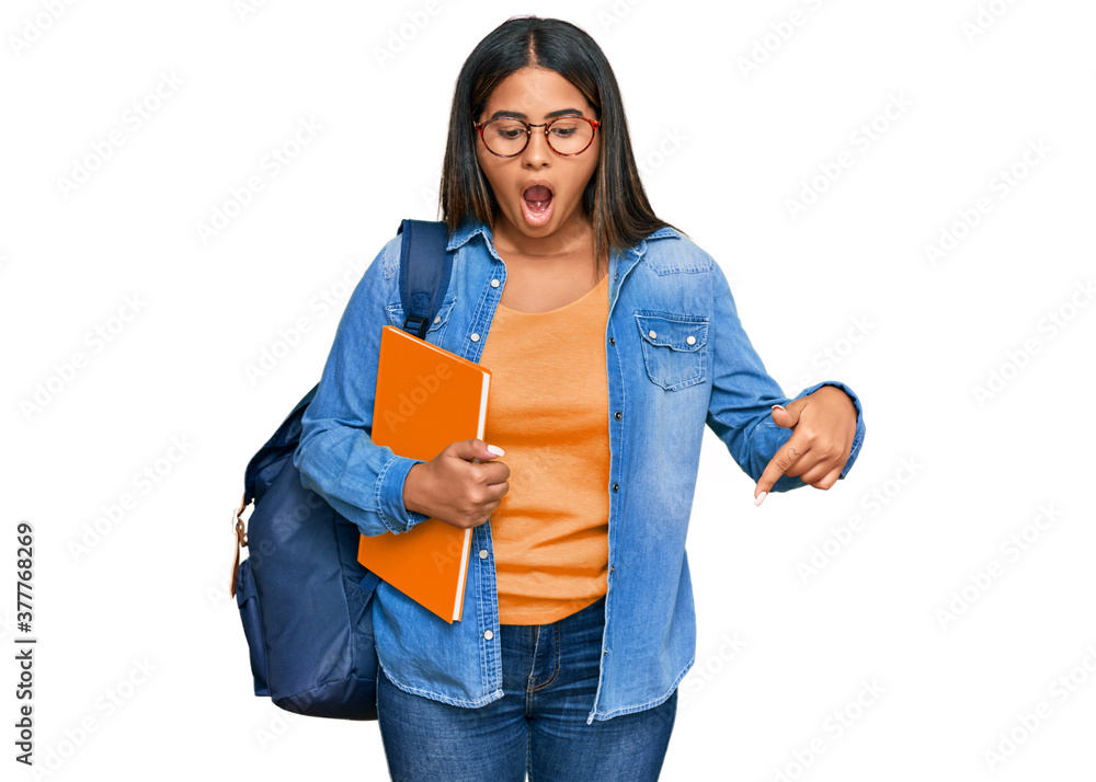 Young latin girl wearing student backpack and holding books pointing down with fingers showing advertisement, surprised face and open mouth Young latin girl wearing student backpack and holding books pointing down with fingers showing advertisement, surprised face and open mouth