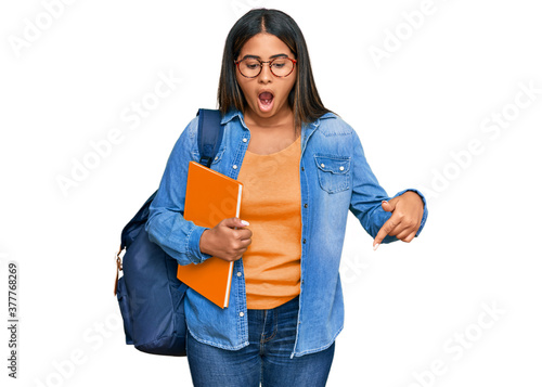 Young latin girl wearing student backpack and holding books pointing down with fingers showing advertisement  surprised face and open mouth