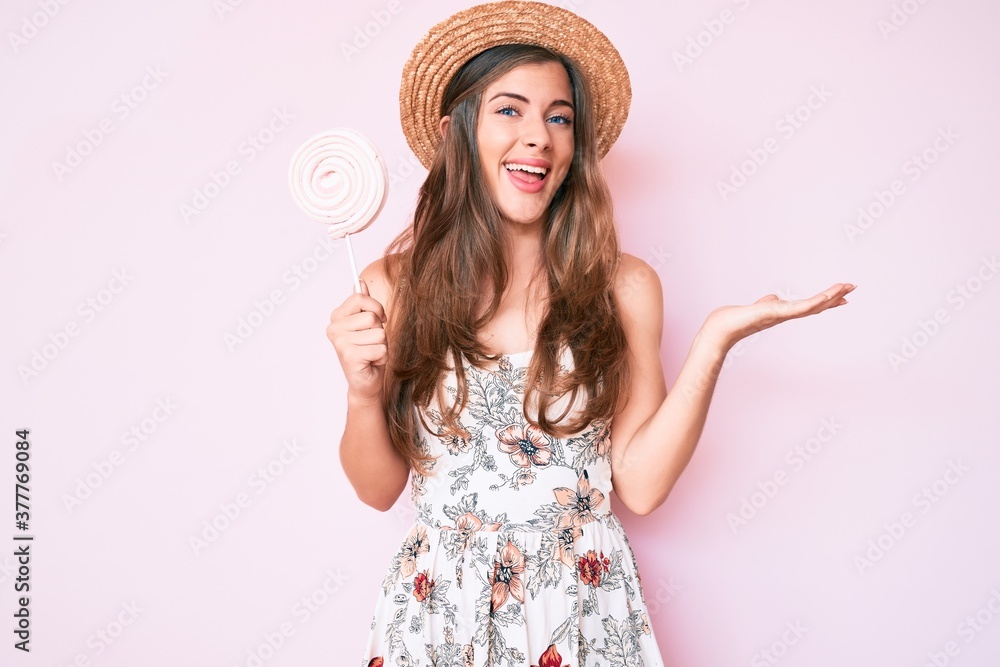Beautiful young caucasian woman wearing summer hat and lollipop celebrating victory with happy smile and winner expression with raised hands