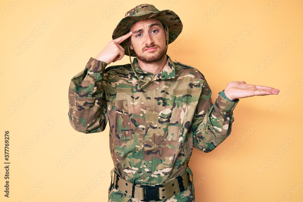 Young caucasian man wearing camouflage army uniform confused and annoyed with open palm showing copy space and pointing finger to forehead. think about it. Young caucasian man wearing camouflage army uniform confused and annoyed with open palm showing copy space and pointing finger to forehead. think about it.