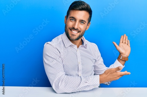 Handsome hispanic man wearing business clothes sitting on the table inviting to enter smiling natural with open hand photo