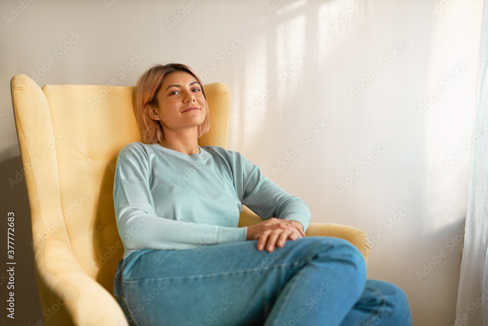 Cute carefree young Caucasian female wearing casual clothes sitting in yellow armchair against white curtain with copy space for your advertisement, having relaxed happy facial expression, smiling Cute carefree young Caucasian female wearing casual clothes sitting in yellow armchair against white curtain with copy space for your advertisement, having relaxed happy facial expression, smiling
