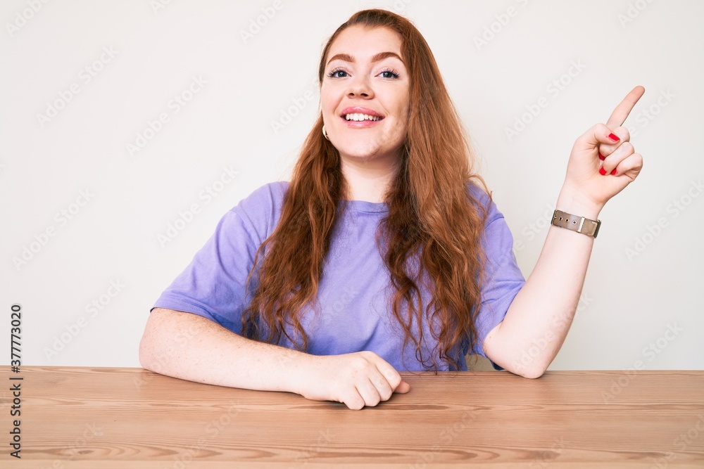 Young redhead woman wearing casual clothes sitting on the table smiling happy pointing with hand and finger to the side Young redhead woman wearing casual clothes sitting on the table smiling happy pointing with hand and finger to the side