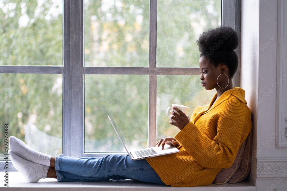 Φωτογραφία Focused African American woman student with afro hairstyle wear yellow cardigan, sitting on windowsill, working doing remote job on laptop, preparing for an exam, holding mug coffee or tea