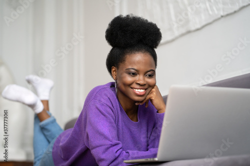 Smiling African American millennial woman with afro hairstyle wear purple sweater lying on sofa, resting, 
looking at camera webcam and talking on a video call or skype with friends, watching movie.  photo